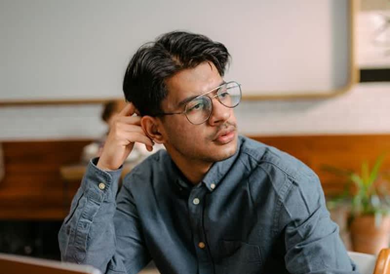 Ein Mann in einem blauen Hemd sitzt an einem Tisch und stützt seinen Kopf mit einer Hand ab. Der Hintergrund zeigt eine Wand mit Holzverkleidung und einem Regal.