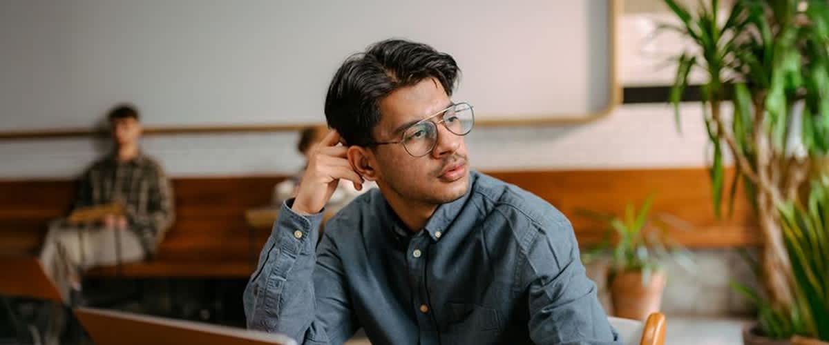 Ein Mann in einem blauen Hemd sitzt an einem Tisch und stützt seinen Kopf mit einer Hand ab. Der Hintergrund zeigt eine Wand mit Holzverkleidung und einem Regal.