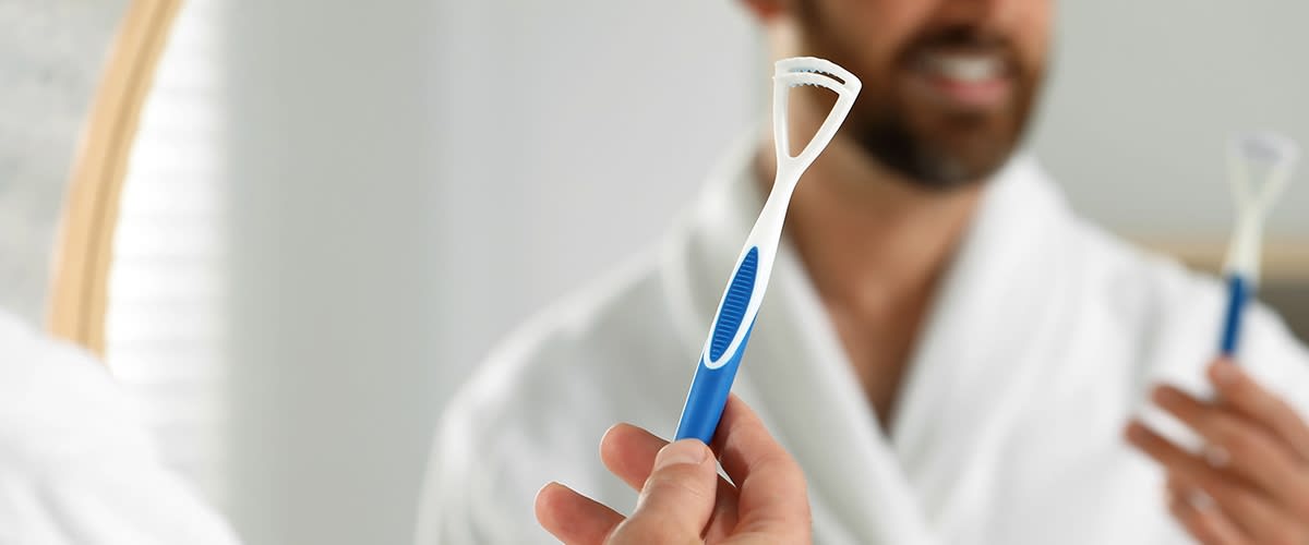 Eine Person in einem weißen Bademantel hält eine blaue Zungenreiniger mit weißen Borsten in der Hand