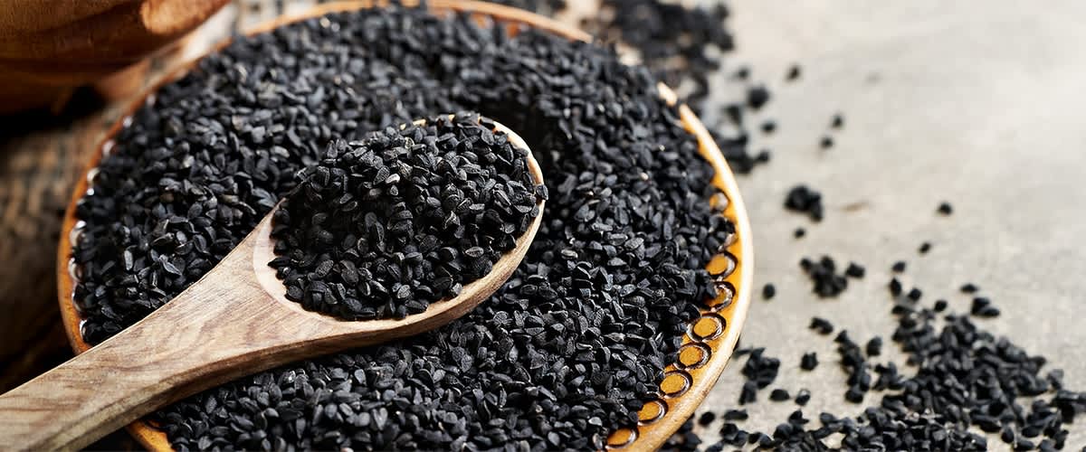 Schwarzkümmel liegt auf einem Holzlöffel und ist teilweise auf einer grauen Oberfläche verstreut