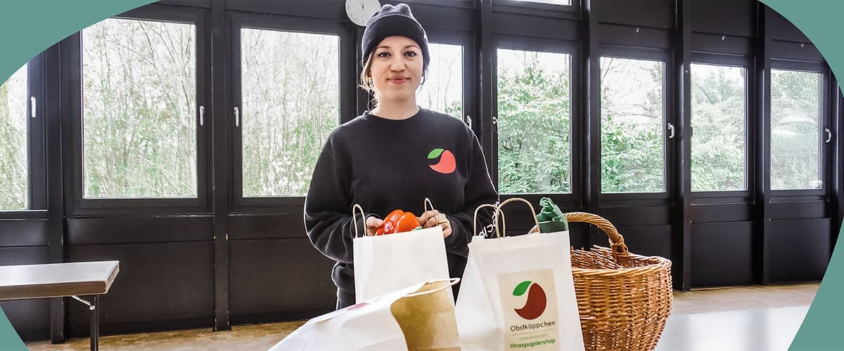 Carina Raddatz in einem schwarzen Pullover mit einem roten Apfel-Logo steht vor großen Fenstern und hält eine Papiertüte mit roten Äpfeln, daneben weitere Papiertüten mit dem Logo von Obstkäppchen und ein Korb