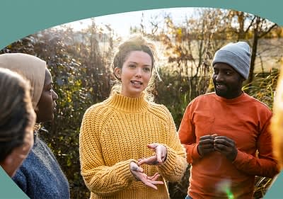 Eine Person in einem gelben Strickpullover gestikuliert mit den Händen, während eine andere Person in einem orangefarbenen Pullover und grauer Mütze aufmerksam zuhört, beide stehen im Freien vor einem Hintergrund aus Büschen und Bäumen