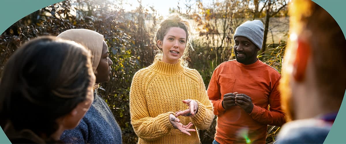Eine Person in einem gelben Strickpullover gestikuliert mit den Händen, während eine andere Person in einem orangefarbenen Pullover und grauer Mütze aufmerksam zuhört, beide stehen im Freien vor einem Hintergrund aus Büschen und Bäumen