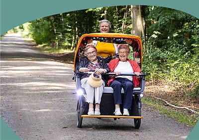 Zwei ältere Personen sitzen in einem gelben Fahrrad-Rikscha auf einem Waldweg, die Frau links trägt eine Brille und hält einen Strohhut in der Hand, während die Person rechts eine rote Jacke trägt
