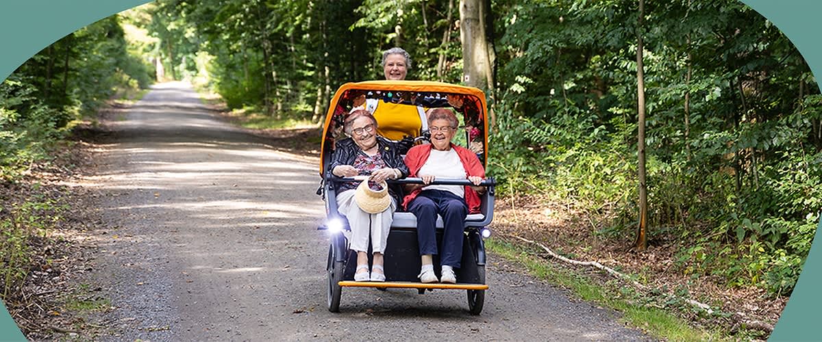 Zwei ältere Personen sitzen in einem gelben Fahrrad-Rikscha auf einem Waldweg, die Frau links trägt eine Brille und hält einen Strohhut in der Hand, während die Person rechts eine rote Jacke trägt