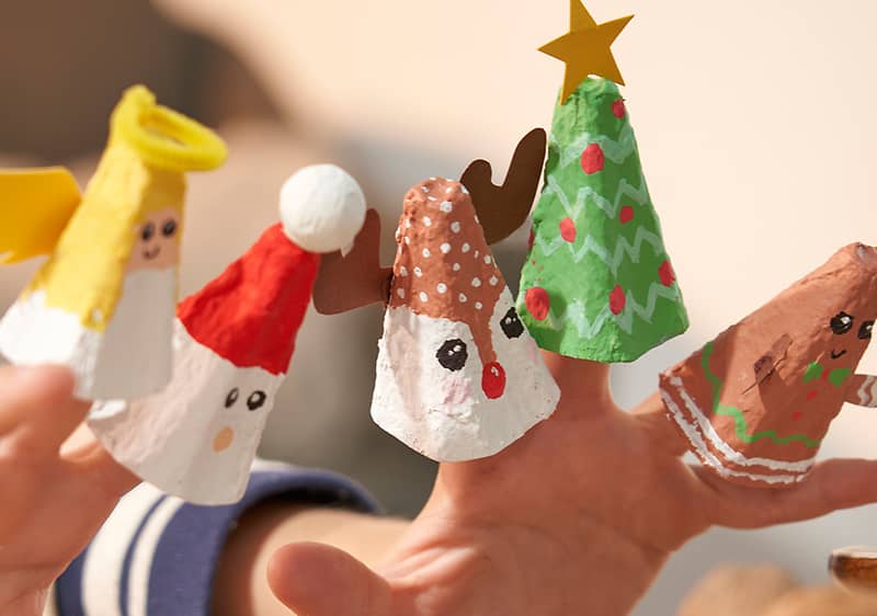 Vier Fingerpuppen aus Eierkartons, die weihnachtliche Figuren darstellen: ein Engel mit gelbem Heiligenschein, ein Weihnachtsmann mit roter Mütze, ein Rentier mit braunen Geweihen und eine grüne Tanne mit gelbem Stern