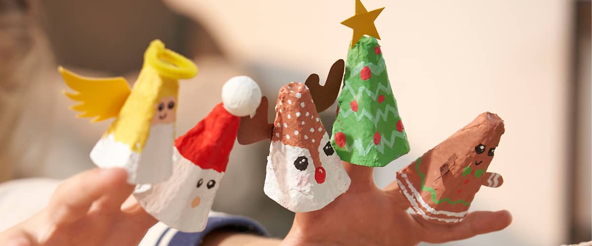 Vier Fingerpuppen aus Eierkartons, die weihnachtliche Figuren darstellen: ein Engel mit gelbem Heiligenschein, ein Weihnachtsmann mit roter Mütze, ein Rentier mit braunen Geweihen und eine grüne Tanne mit gelbem Stern