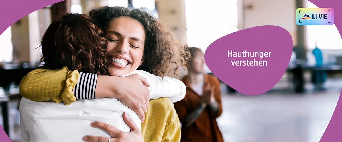 Zwei Personen umarmen sich herzlich, eine trägt ein gestreiftes Hemd, die andere eine gelbe Jacke. Rechts im Bild steht der Text „Hauthunger verstehen“ in einem lila Bereich. Oben rechts ist ein kleines Logo mit der Aufschrift „dmLIVE“