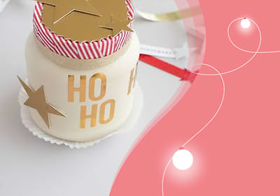 Ein weißes Glas mit rotem und weißem gestreiftem Deckel, goldenen Sternen und der Aufschrift „HO HO“ steht auf einem weißen Untersetzer, daneben eine rote und weiße Schleife auf rosa Hintergrund mit weißen Lichtpunkten