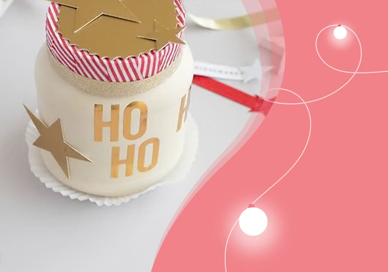 Ein weißes Glas mit rotem und weißem gestreiftem Deckel, goldenen Sternen und der Aufschrift „HO HO“ steht auf einem weißen Untersetzer, daneben eine rote und weiße Schleife auf rosa Hintergrund mit weißen Lichtpunkten