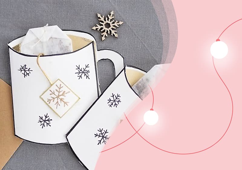 Eine Illustration zeigt eine weiße Teetasse mit schwarzen Schneeflockenmustern und einem Teebeutel mit Schneeflockenanhänger auf einem grauen Hintergrund, daneben eine rosa Fläche mit weißen Lichterketten