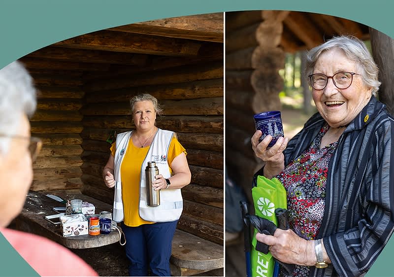 Zwei ältere Personen stehen vor einer Holzhütte, eine Person hält eine Thermoskanne und eine Tasse, die andere Person hält ein Glas und eine grüne Tasche.