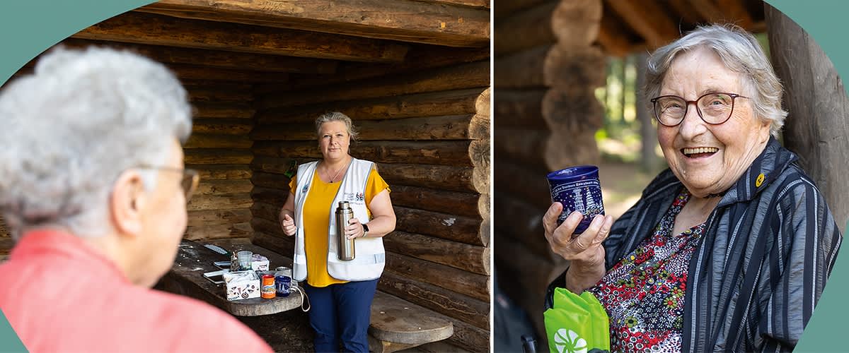 Zwei ältere Personen stehen vor einer Holzhütte, eine Person hält eine Thermoskanne und eine Tasse, die andere Person hält ein Glas und eine grüne Tasche.