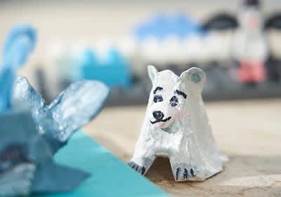 Ein weißer, handbemalter Eisbär aus Papiermaché mit schwarzen Augen und einem freundlichen Gesichtsausdruck steht auf einem Tisch, im Hintergrund sind unscharfe blaue und graue Objekte zu sehen