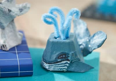 Ein aus EIerkarton gebastelter Wal mit blauen Pfeifenreinigern als Wasserfontäne auf einem türkisfarbenen Untergrund