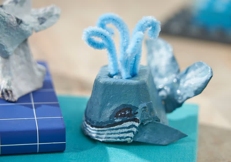 Ein aus EIerkarton gebastelter Wal mit blauen Pfeifenreinigern als Wasserfontäne auf einem türkisfarbenen Untergrund