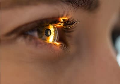 Nahaufnahme eines menschlichen Auges, das von einem hellen Lichtstrahl beleuchtet wird, wodurch die Iris in einem goldenen Farbton erscheint und die Wimpern deutlich hervorgehoben werden