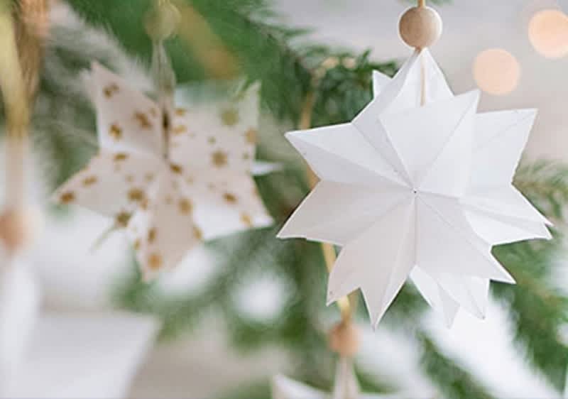 Zwei weiße Origami-Sterne hängen an einem grünen Tannenzweig, der hintere Stern hat goldene Punkte