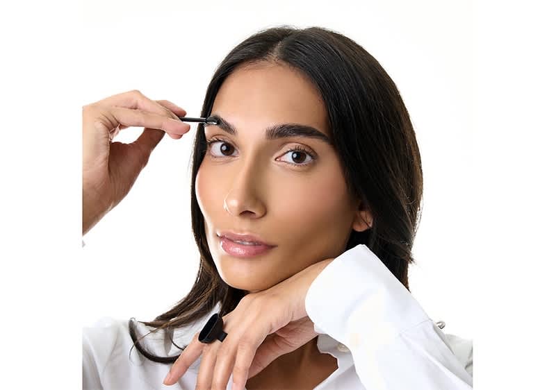 Eine Person in weißem Hemd hält einen schwarzen Eyeliner in der rechten Hand und führt ihn zur Augenbraue