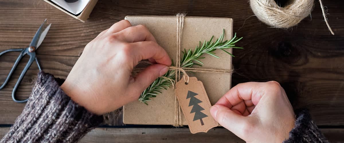 Zwei Hände binden eine Kordel um ein braunes Geschenk, das mit einem Zweig und einem Etikett mit einem Tannenbaum verziert ist