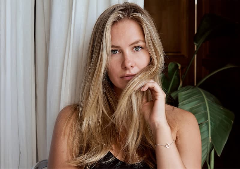 Eine Person mit langen, blonden Haaren trägt ein schwarzes Oberteil und hält eine Haarsträhne mit der Hand