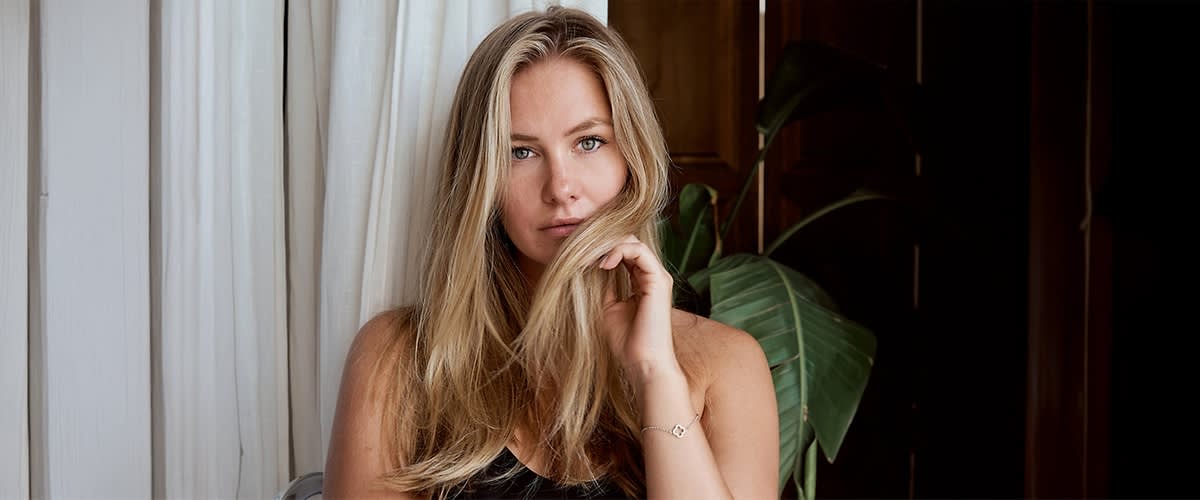 Eine Person mit langen, blonden Haaren trägt ein schwarzes Oberteil und hält eine Haarsträhne mit der Hand