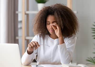 Eine Person mit lockigem Haar sitzt an einem Tisch, hält eine Brille in der Hand und arbeitet an einem Laptop, im Hintergrund sind Regale mit Pflanzen sichtbar