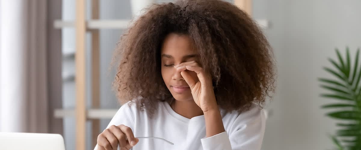 Eine Person mit lockigem Haar sitzt an einem Tisch, hält eine Brille in der Hand und arbeitet an einem Laptop, im Hintergrund sind Regale mit Pflanzen sichtbar