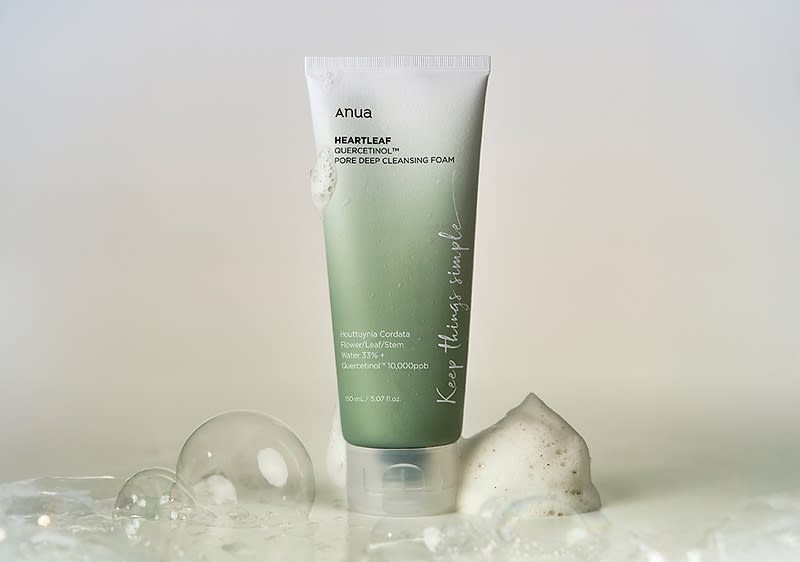 Eine grüne Tube mit der Aufschrift „Anua Heartleaf 70% Pore Deep Cleansing Foam“ steht auf einer nassen Oberfläche, umgeben von Schaum und Wasserblasen