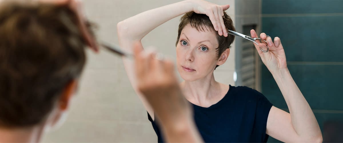 Eine Frau in einem dunkelblauen T-Shirt schneidet sich mit einer Schere die Haare vor einem Spiegel.
