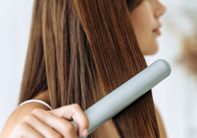 Eine Person glättet ihr langes, braunes Haar mit einem silbernen Haarglätter