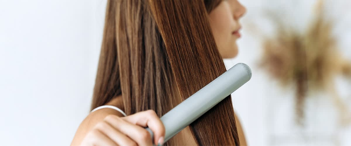 Eine Person glättet ihr langes, braunes Haar mit einem silbernen Haarglätter