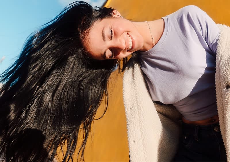 Eine Person mit langen, dunklen Haaren trägt ein helles Oberteil und eine flauschige Jacke, der Hintergrund ist orangefarben und blau