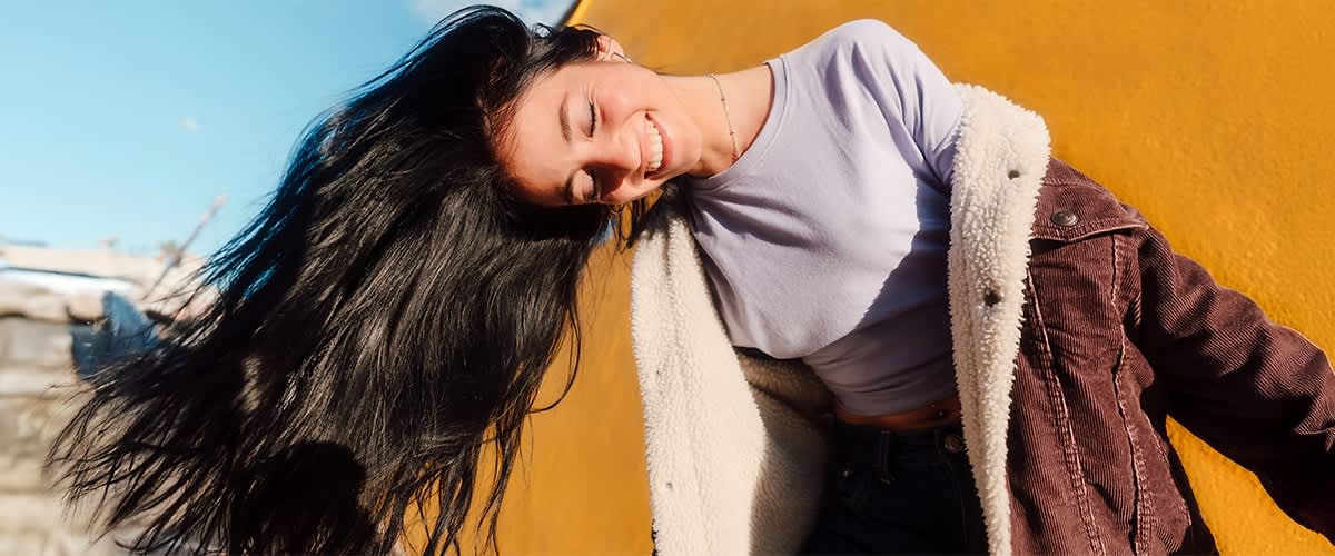 Eine Person mit langen, dunklen Haaren trägt ein helles Oberteil und eine flauschige Jacke, der Hintergrund ist orangefarben und blau