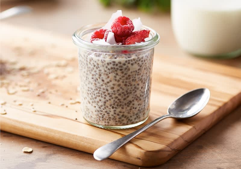 Ein Glas mit Chia-Pudding, garniert mit Himbeeren und Kokosflocken, steht auf einem Holzbrett neben einem Löffel