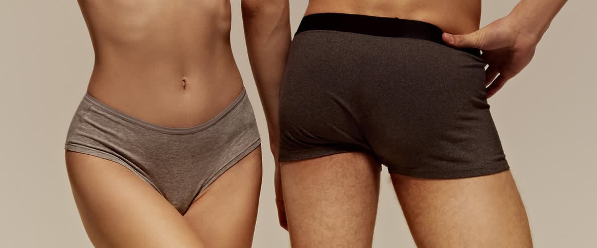 Nahaufnahmen der Becken- und Oberschenkelpartie von zwei Personen, links eine Person mit grauen Slip-Unterhosen, rechts eine Person mit dunkelgrauen Boxershorts, beide stehend, rechte Hand der rechten Person am Hüftbund der Boxershorts, neutraler einfarbiger Hintergrund