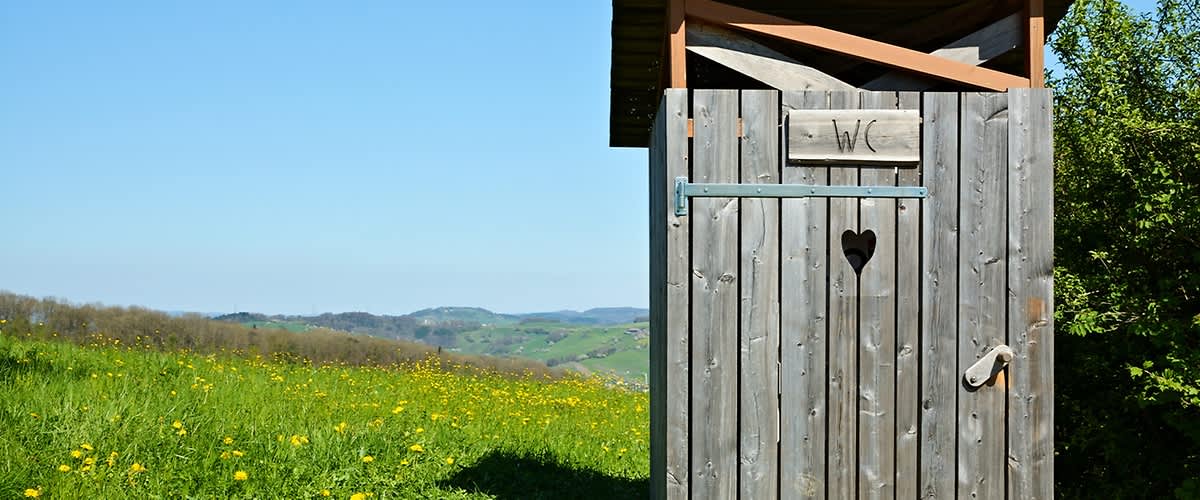Eine einfache Holzhütte als Außen-Toilette mit ausgeschnittenem Herz im Türblatt und der Aufschrift „WC“ über der Tür, stehend auf einer sonnigen Wiese mit gelben Blumen und hügeliger Landschaft im Hintergrund