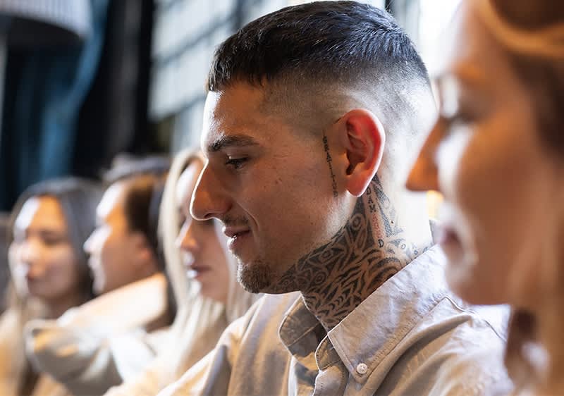 Ein Mann mit tätowiertem Hals und Crew Cut trägt ein helles Hemd. Im Hintergrund sind weitere Personen unscharf zu erkennen.