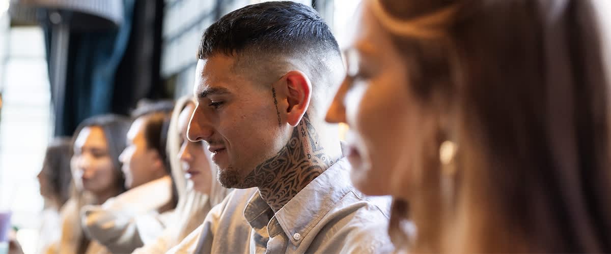 Ein Mann mit tätowiertem Hals und Crew Cut trägt ein helles Hemd. Im Hintergrund sind weitere Personen unscharf zu erkennen.
