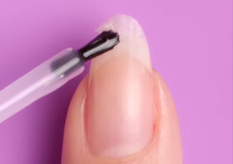 Ein Pinsel trägt schwarzen Nagellack auf einen langen, natürlichen Fingernagel auf, vor einem lilafarbenen Hintergrund