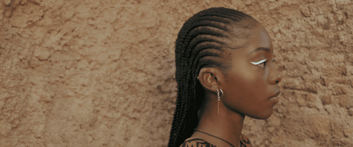 Eine Person mit Cornrows und einem Ohrring steht vor einer Wand aus grobem Stein.