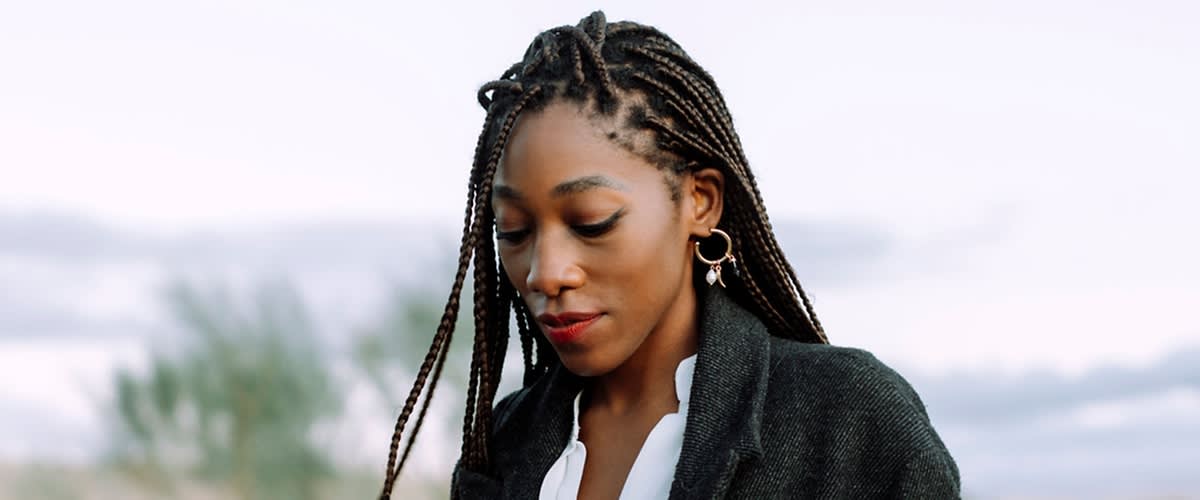Eine Person mit langen, geflochtenen Haaren trägt goldene Ohrringe und einen dunklen Mantel über einem weißen Oberteil