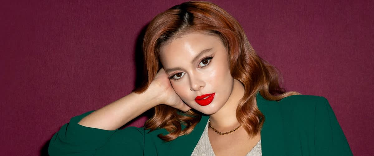 Eine Person mit welligem, kastanienbraunem Haar trägt ein grünes Oberteil und eine goldene Halskette vor einem dunkelroten Hintergrund