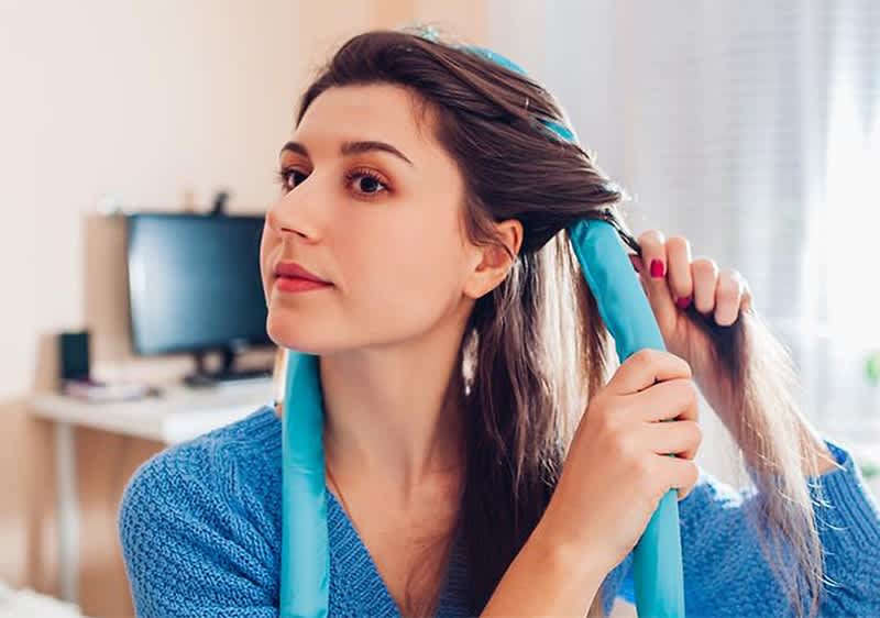 Eine Person mit langen braunen Haaren wickelt ihr Haar um einen blauen Lockenwickler, während sie ein blaues Strickoberteil trägt