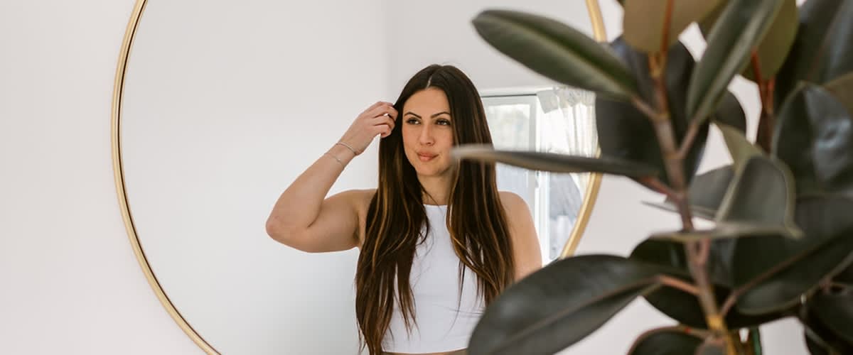 Eine Frau mit langen braunen Haaren steht vor einem runden Spiegel und trägt ein weißes ärmelloses Oberteil, im Vordergrund sind unscharfe Blätter zu sehen