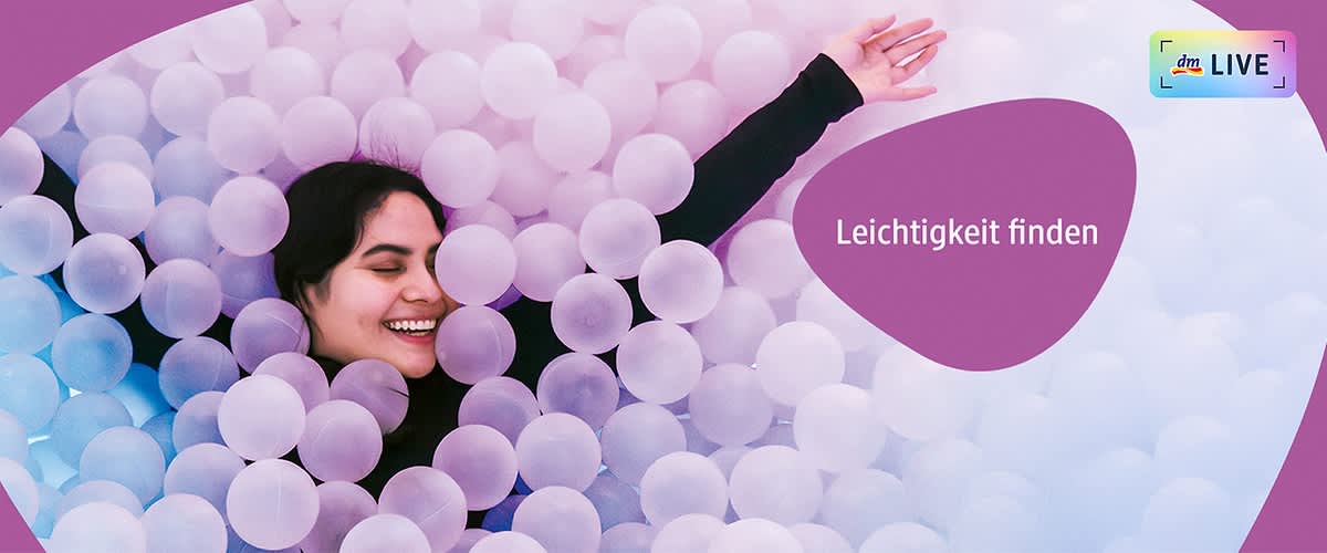 Eine Person liegt in einem Bällebad aus weißen und lilafarbenen Bällen, der Text „Leichtigkeit finden“ ist in weißer Schrift auf einem lilafarbenen Hintergrund zu sehen, oben rechts befindet sich das dmLIVE Logo