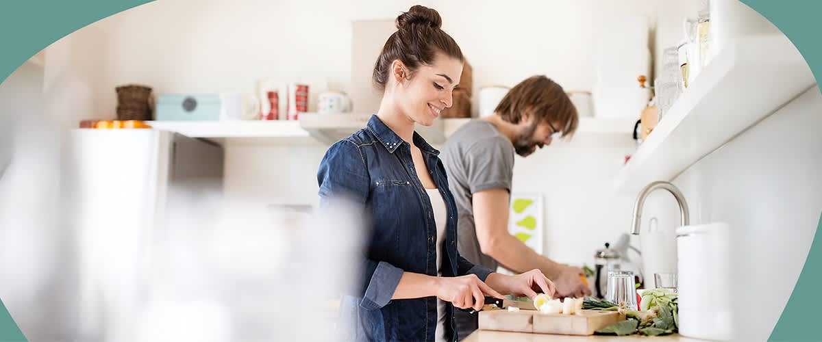 Zwei Personen stehen in einer modernen Küche und schneiden Gemüse auf einem Holzbrett.