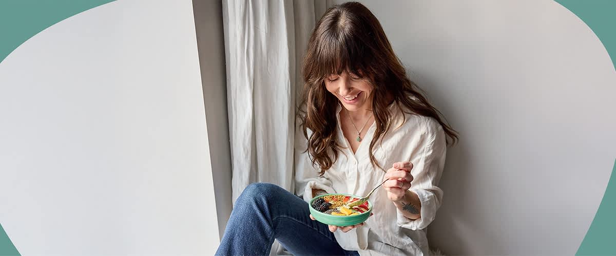 Eine Person in weißer Bluse und blauer Jeans sitzt auf einem Kissen und hält eine Schüssel mit verschiedenen Lebensmitteln, darunter Avocado, Tomaten und Reis