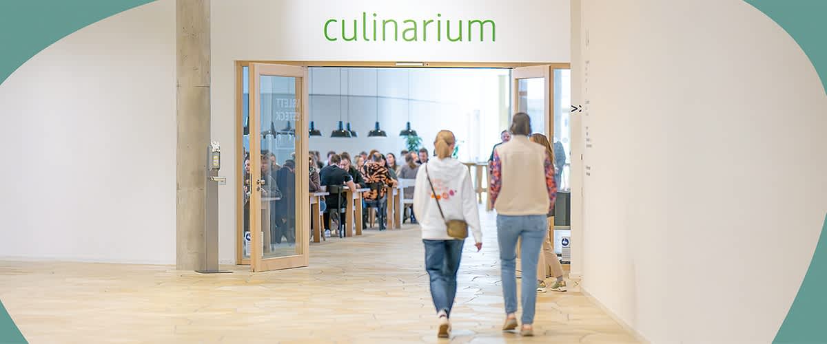 Zwei Personen gehen auf das culinarium zu, in dem viele Menschen bereits an Tischen sitzen und das zubereitete Essen genießen.