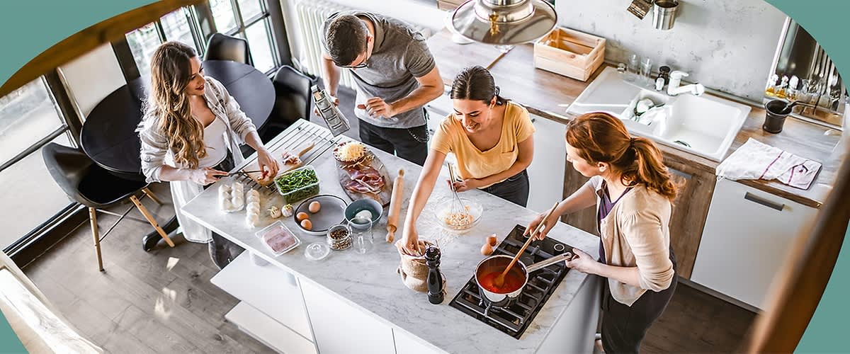 Vier Personen bereiten gemeinsam eine Mahlzeit in einer modernen Küche zu. Auf der Arbeitsfläche liegen verschiedene Zutaten wie Gemüse, Eier und Brot. Eine Person rührt in einem Topf mit roter Sauce auf dem Herd.